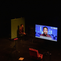 An actor on stage facing a TV Screen.