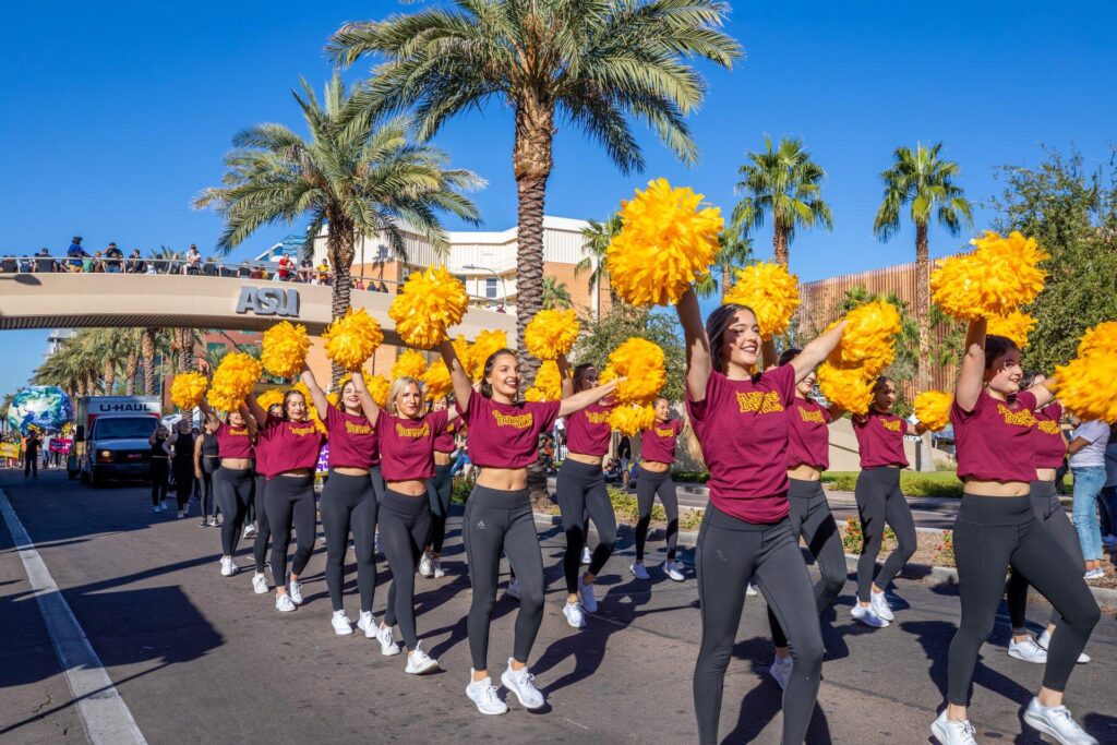 A parade of ASU Cheerleaders with pom poms up in the air marching down the street
