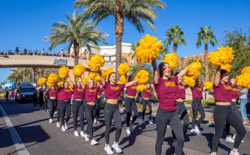 A parade of ASU Cheerleaders with pom poms up in the air marching down the street