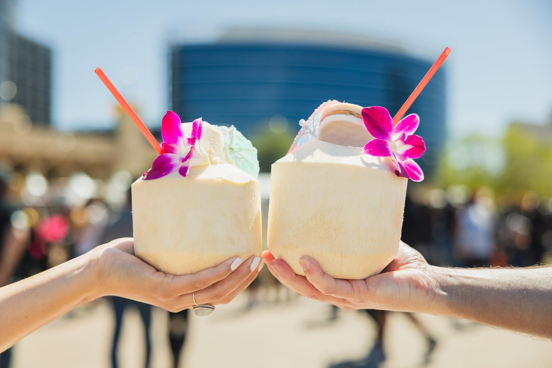 Two hands holding coconuts topped with flowers, umbrellas, and straws at the Arizona Aloha Festival in Tempe.