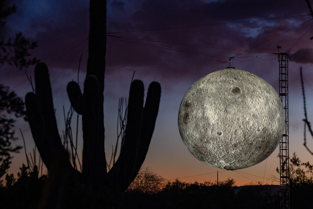 A large moon installation hangs against a desert sunset sky.